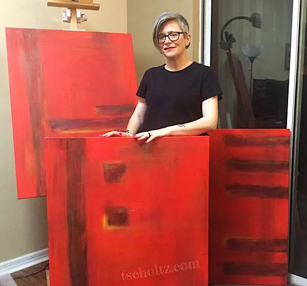 the artist Tes Scholtz shown standing among three large abstract paintings. The paintings are mostly a warm red with other colors throughout.